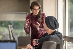 Eine Frau mit einem roten Pullover und langen Haaren steht gestikulierend vor einem interessiert wirkendem Mann - Symbolbild